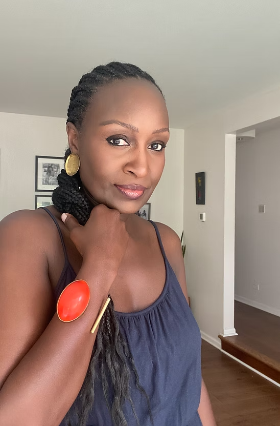 Woman with braided hair wearing a blue top and orange bracelet indoors.